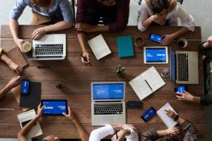 Overhead view of people sitting around a conference table with multiple laptops, tablets and notepads.